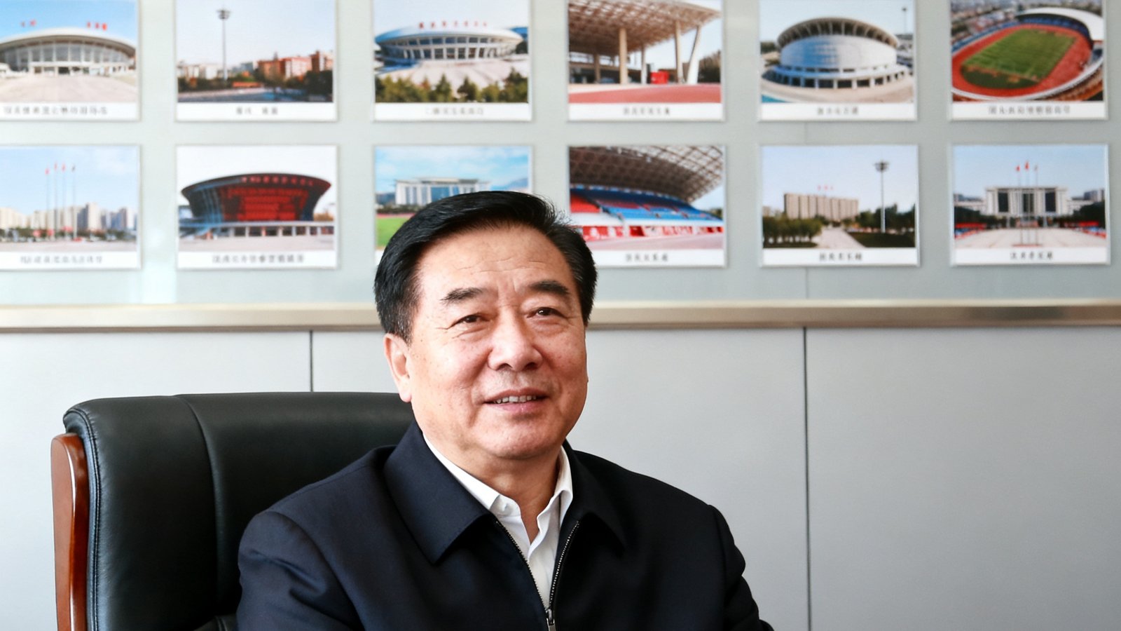 河北省沧州市体育局局长李明辉在办公室接受采访时的正面肖像，背景为沧州体育馆照片墙